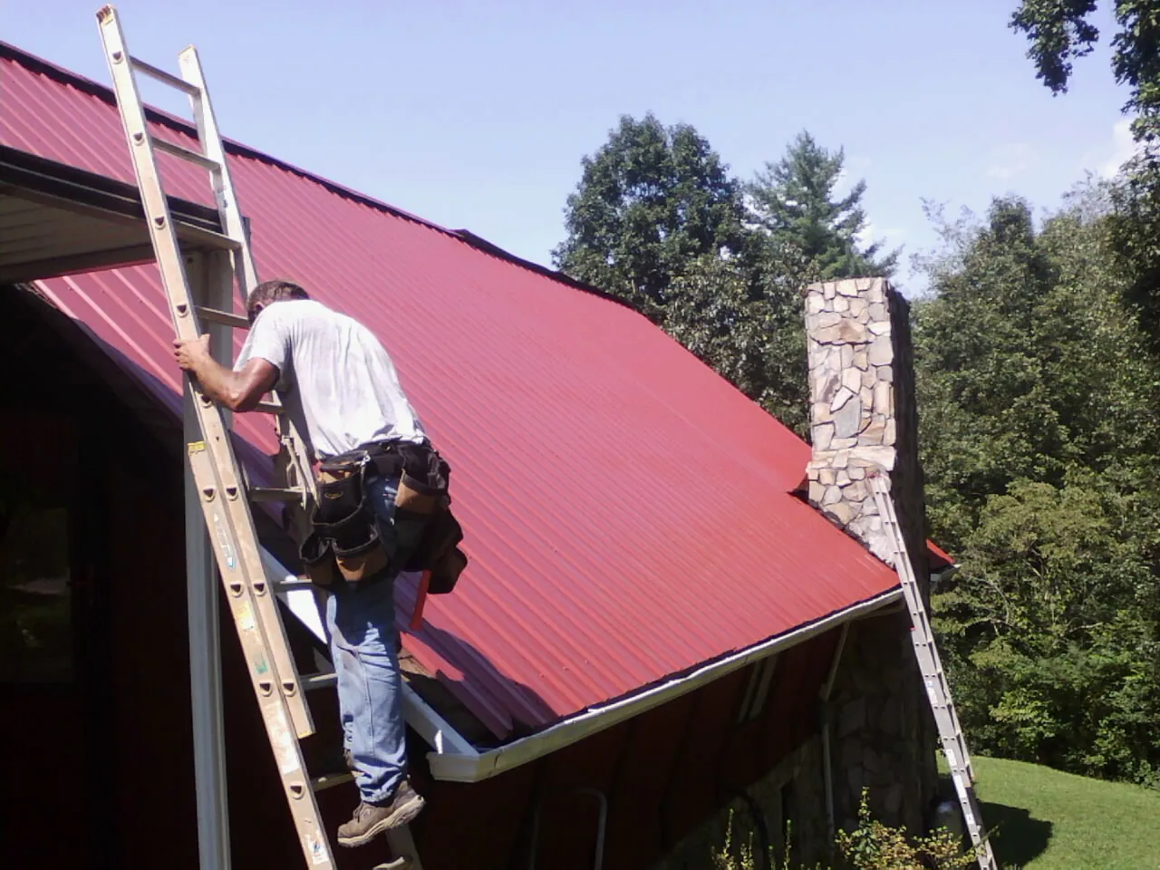 Roofer on a ladder beside a red metal roof and stone chimney — Soffit & Fascia Repair by Roofing Brothers in Modena